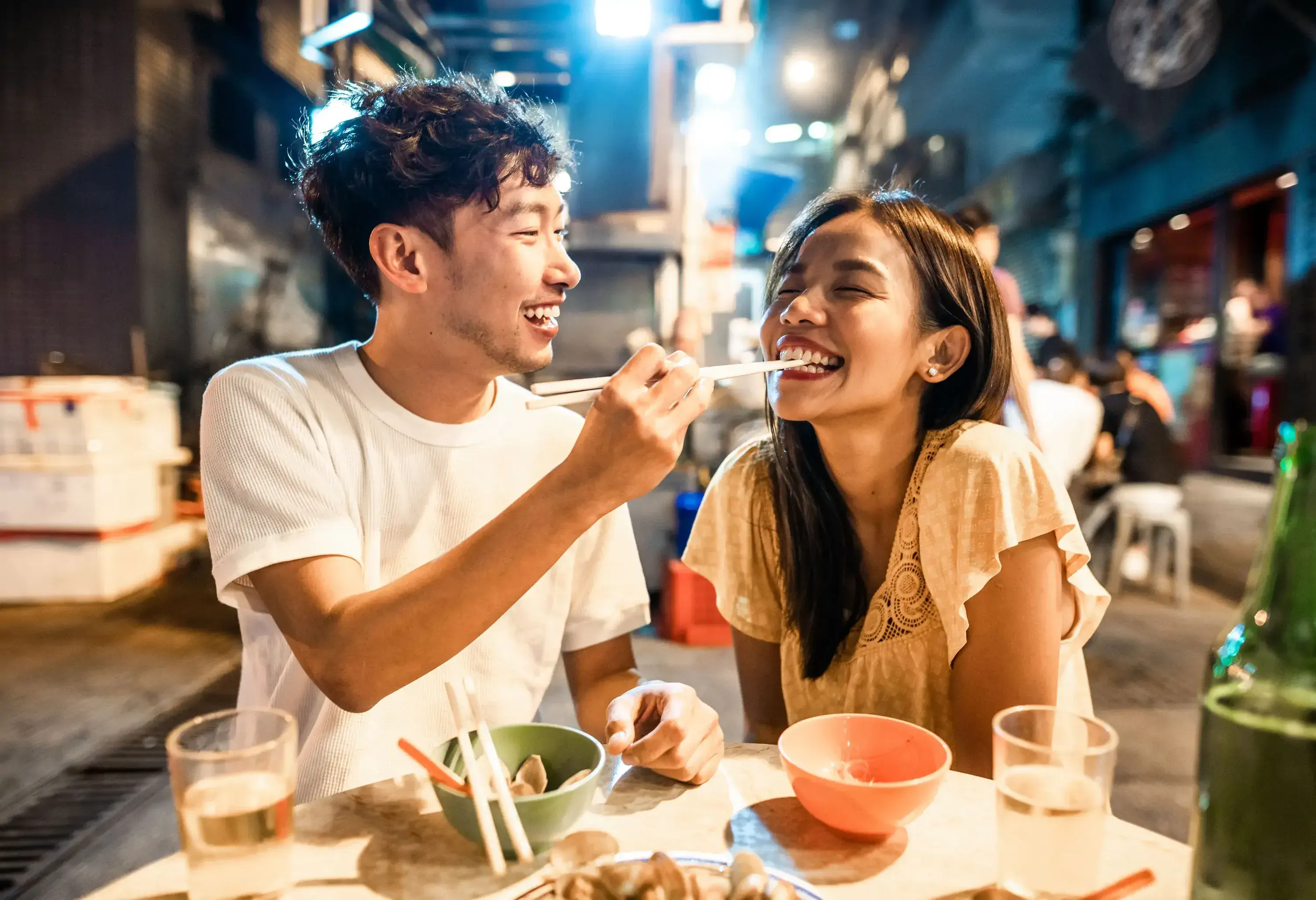 A man and woman smile as he feeds her street food using chopsticks.