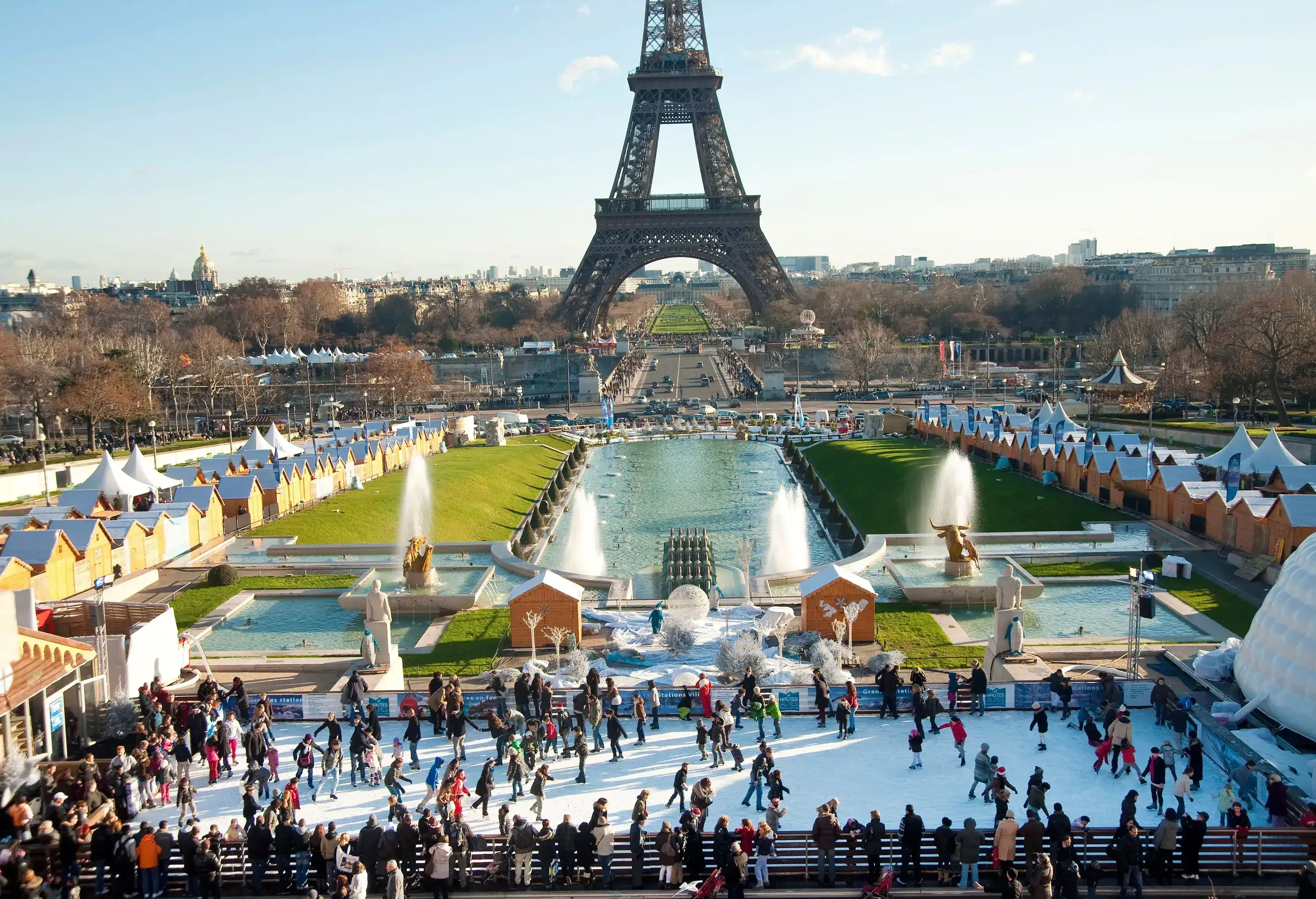 Outdoor ice rink filled with people, set in a park with fountains and a large iron tower in the background.