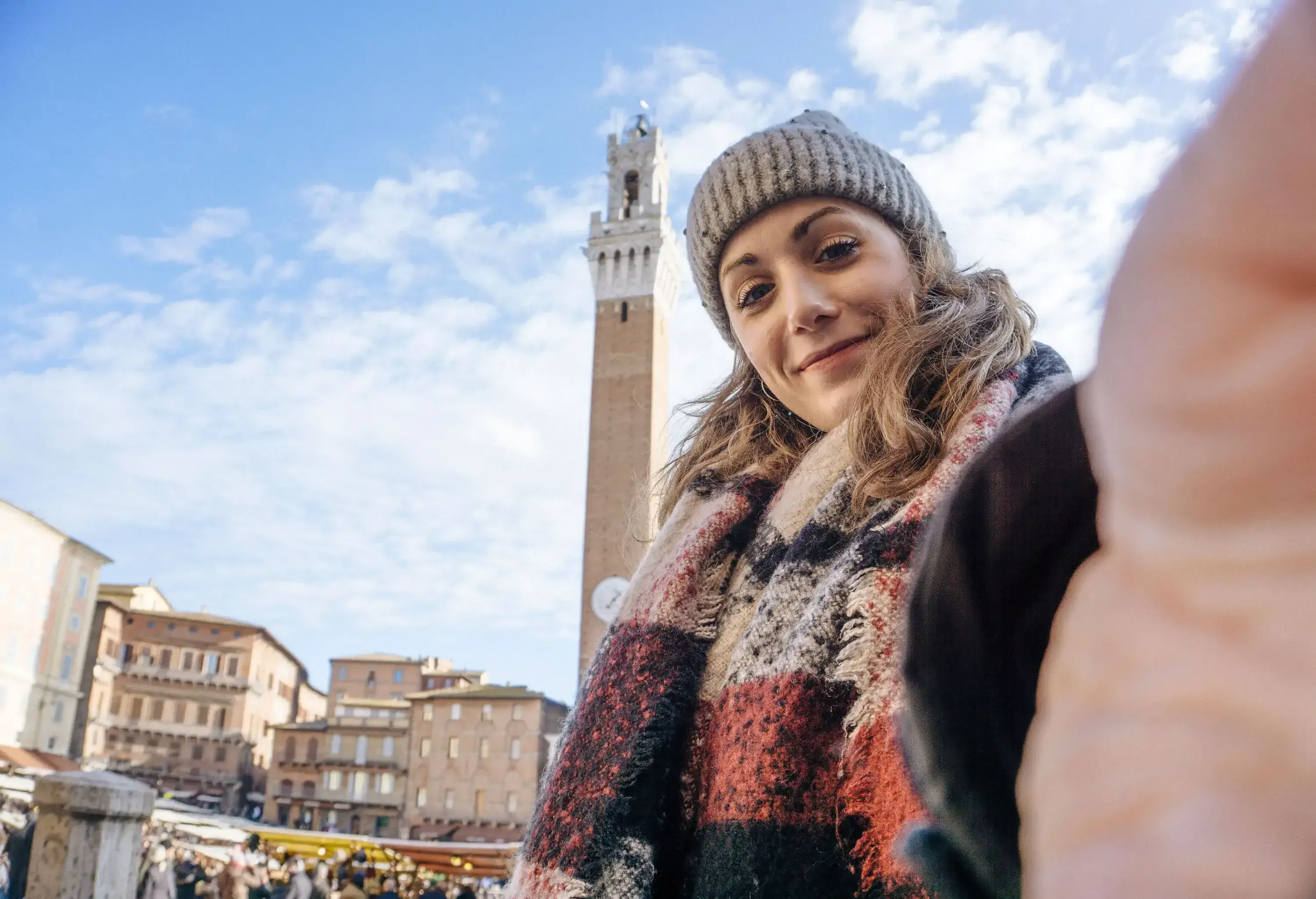 A lady in winter clothes takes a selfie against an imposing tower on a bustling square.