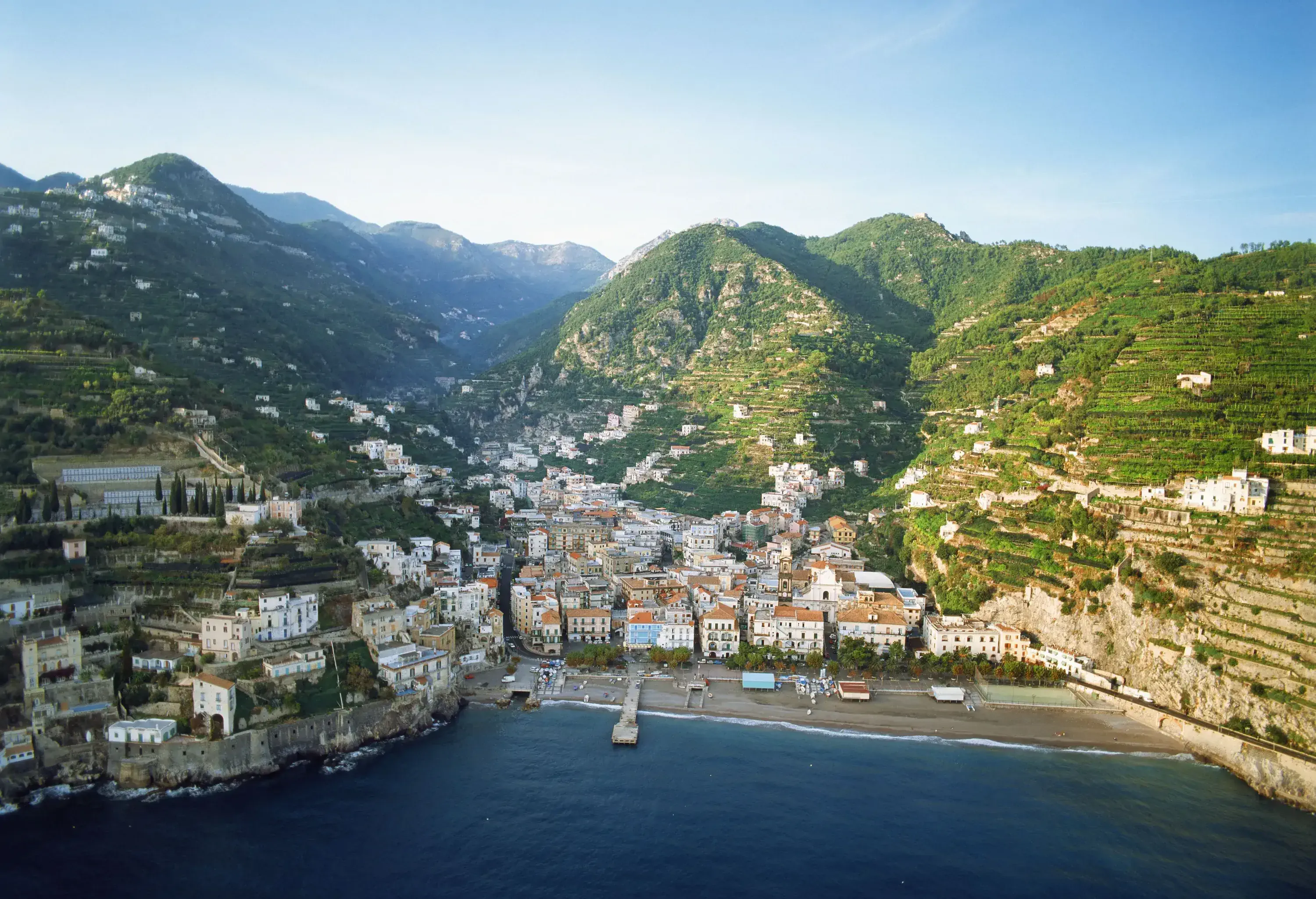 Clifftop houses along a tranquil bay backed by mountains and hillside plantation fields.