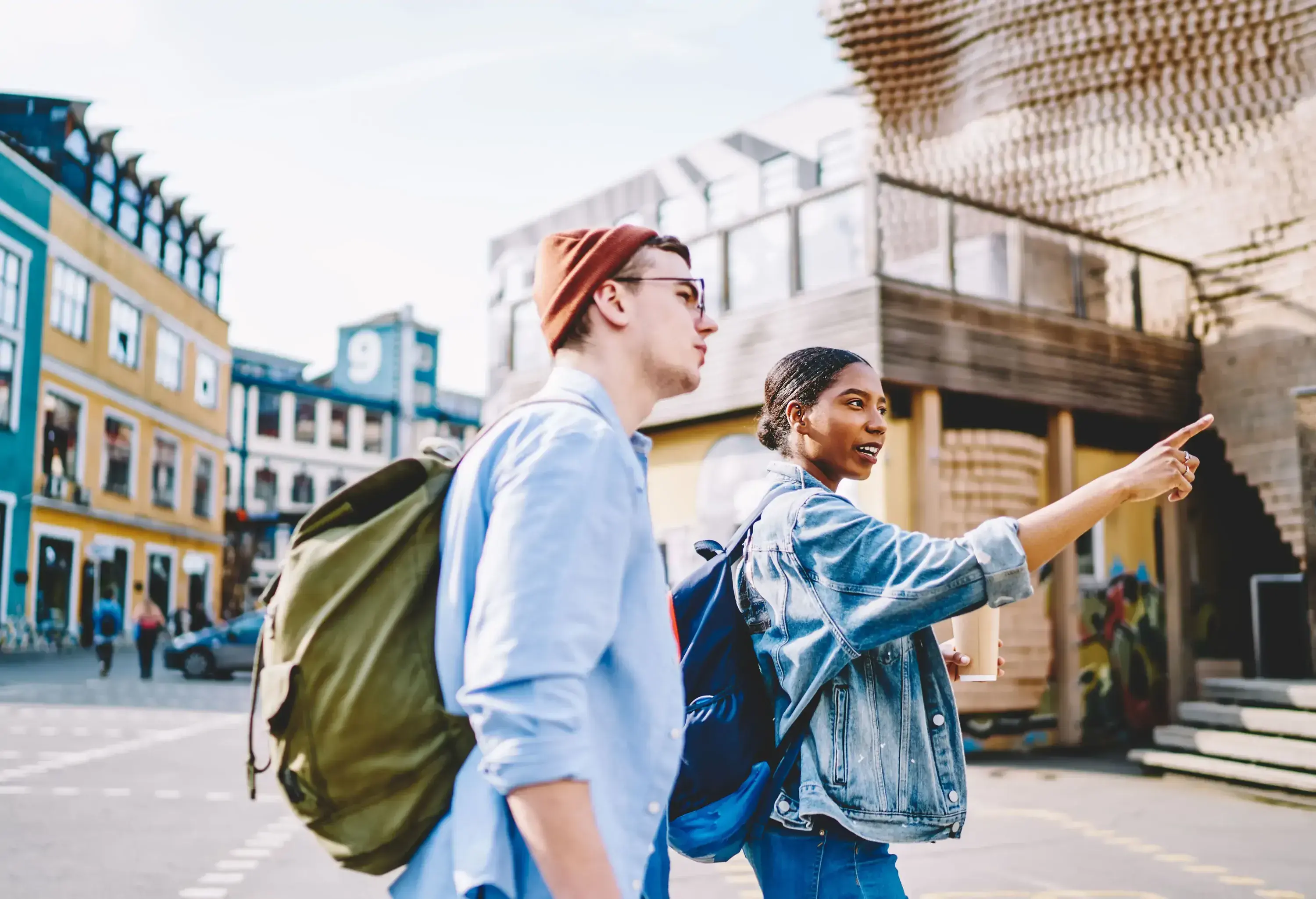 Two friends in casual clothes and backpacks stand on a street, one pointing at something as the other observes.