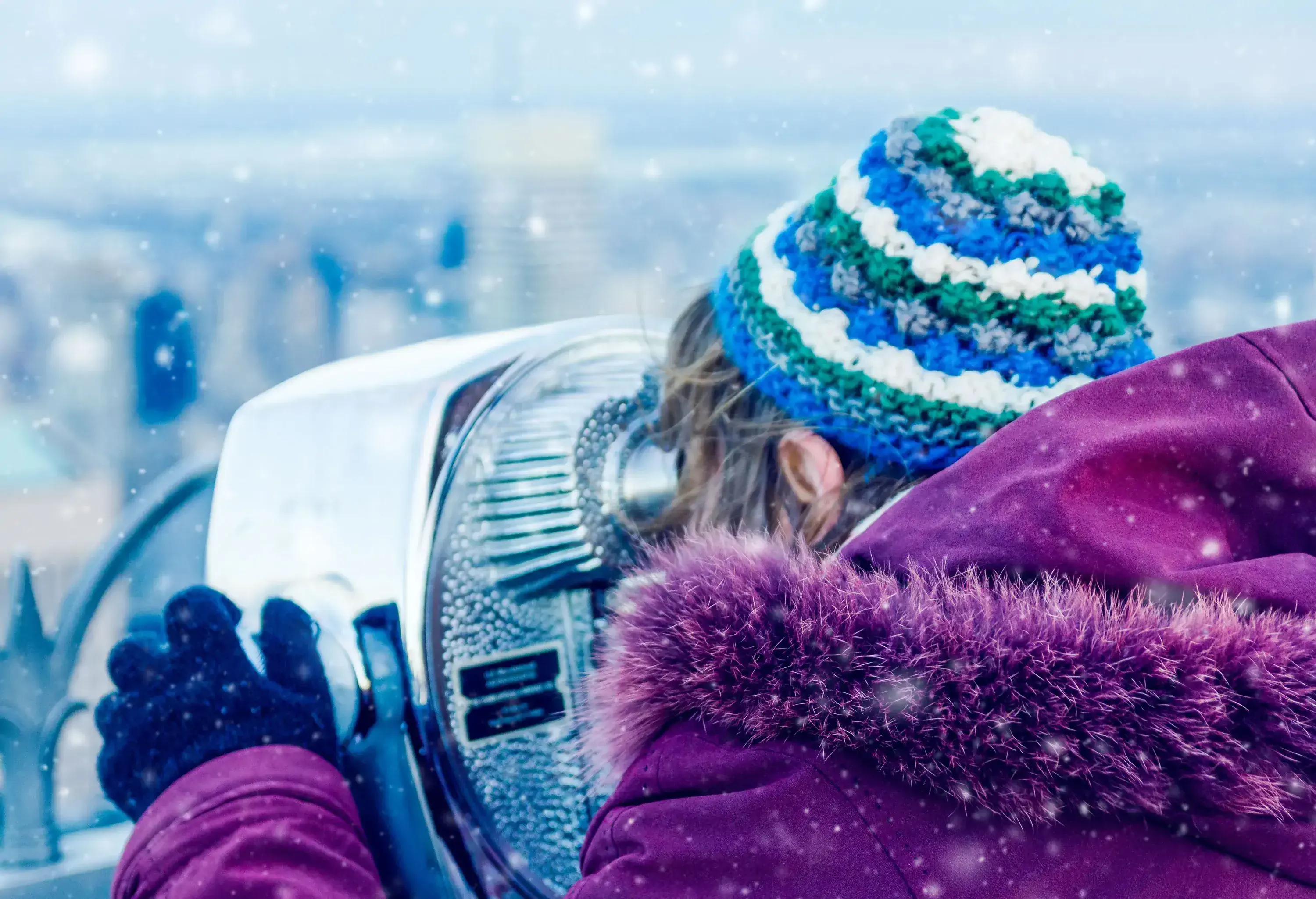 A person in a knitted bonnet, fur coat, and black gloves peers into a coin operated binoculars during snowfall.