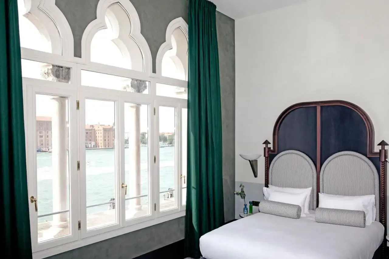Elegant hotel room with a large arched window overlooking a canal. The room features a plush, white bed with a dark, ornate headboard and green curtains.