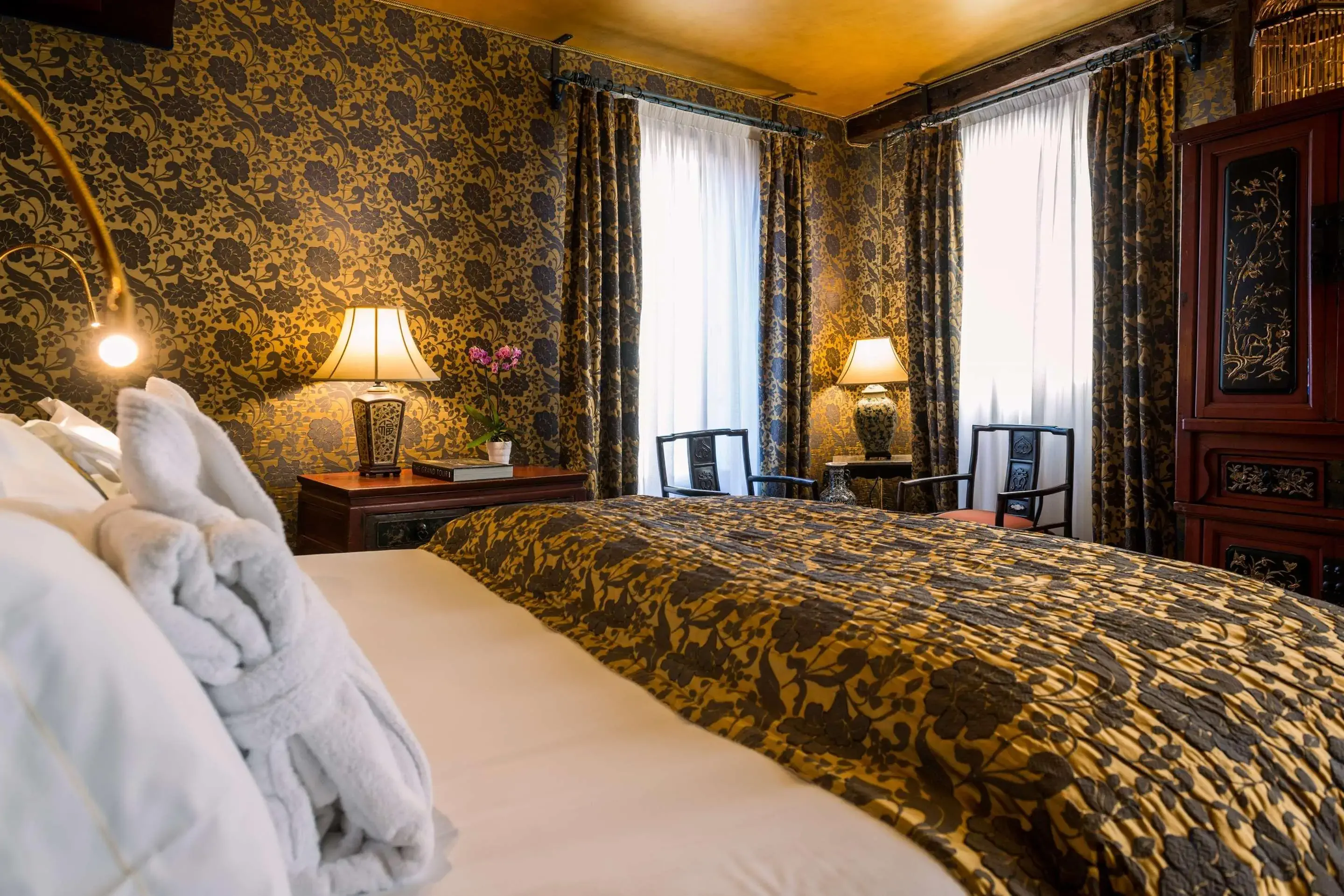 Elegant bedroom with rich floral wallpaper and bedding, soft lighting from lamps, and two tall windows draped in matching curtains. 