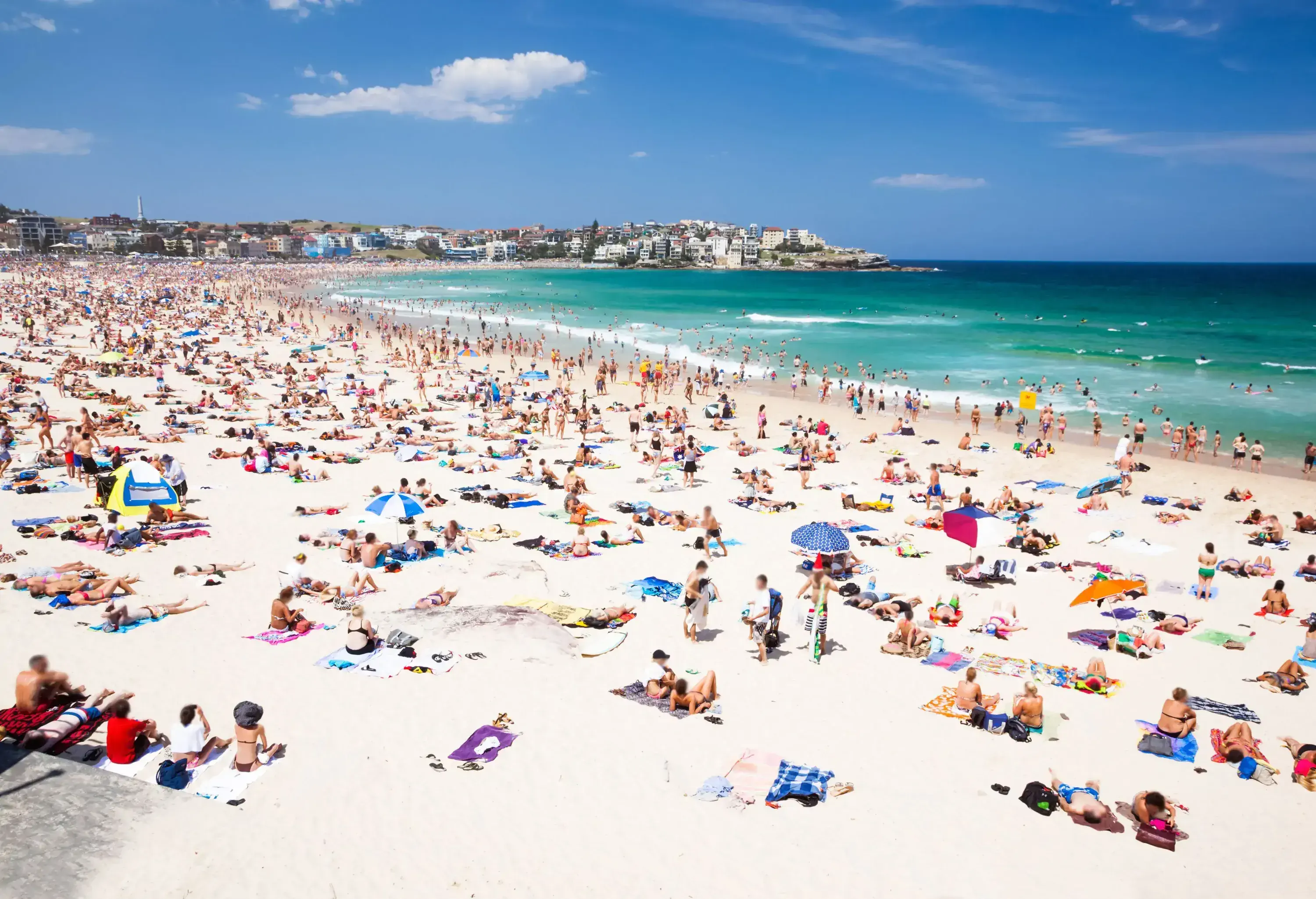 A very crowded beach with people lounging on towels on the sand and swimming in the blue seas.