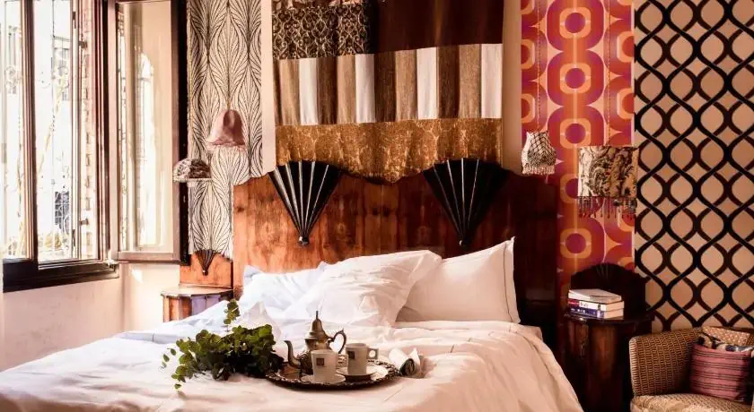 Cozy bedroom with an eclectic mix of patterns. A wooden headboard features diverse fabric panels. A tray with a teapot and cups sits on a rumpled white bedspread. The room is warmly lit through an open window.