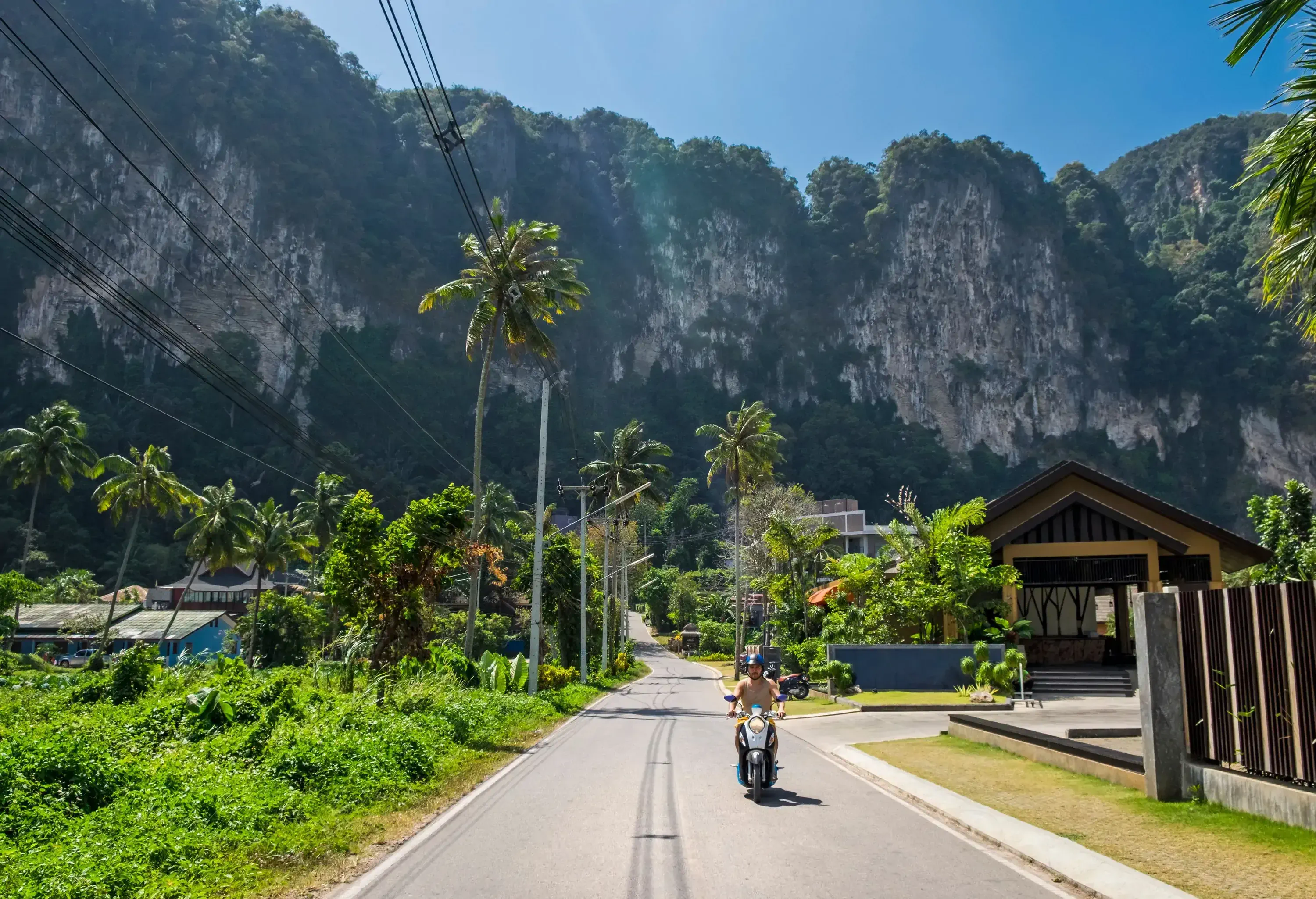 A man cruises effortlessly on his scooter along a beautiful winding road, immersed in verdant tropical scenery, with the imposing presence of a cliff providing a dramatic backdrop in the distance.