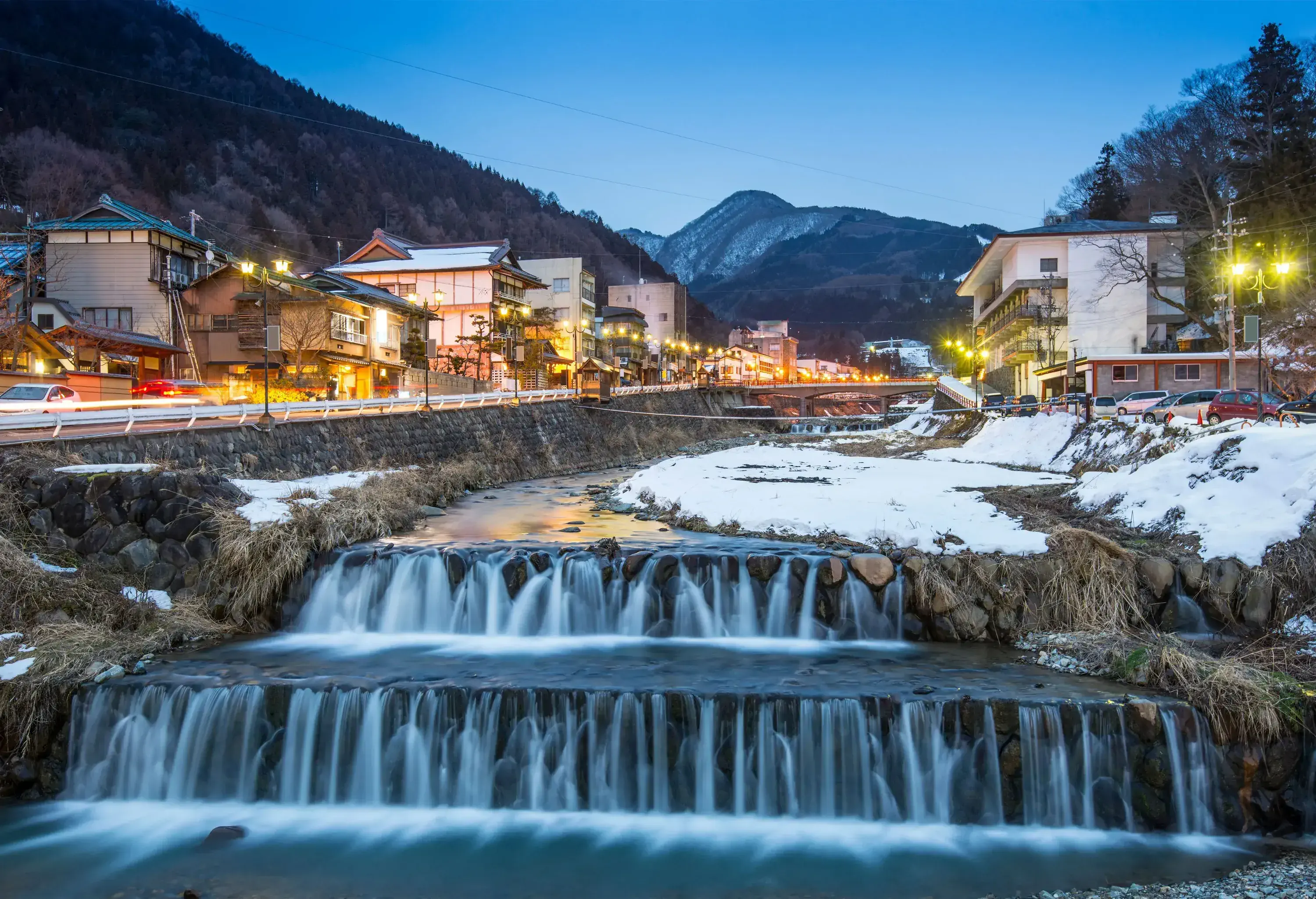 Shibu Onsen - a terraced river lined with the bustling road and buildings beneath the forested hill captured at twilight.