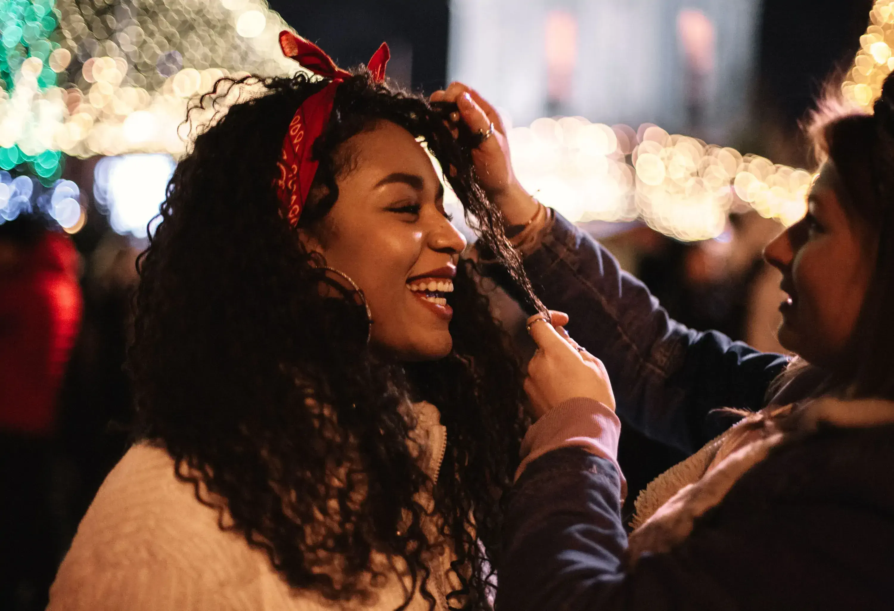 A smiling young woman wearing a red headband looking at another person who touches her hair.