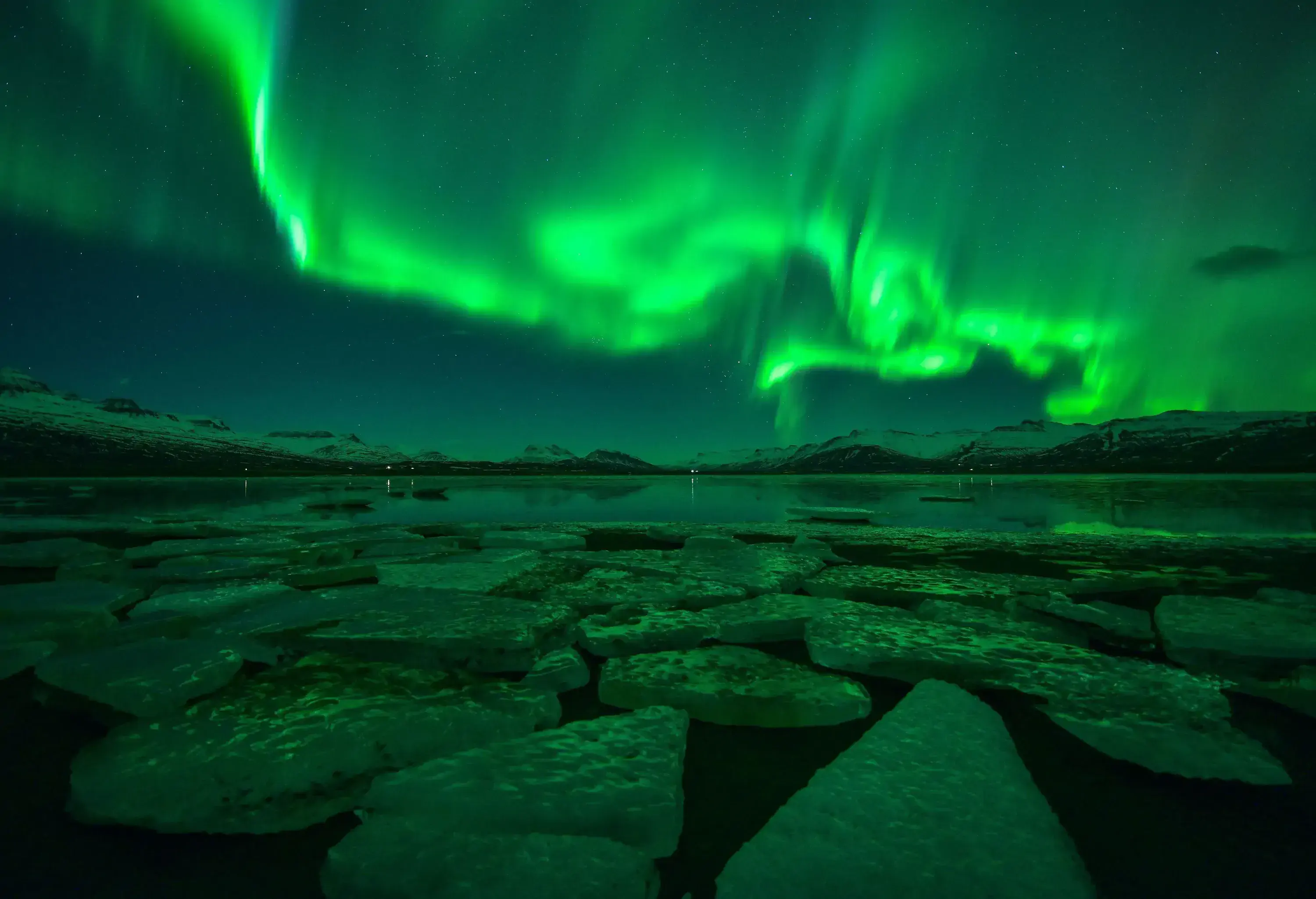 At nighttime, green aurora borealis illuminates the sky above a calm lagoon covered in shards of ice, with snow-covered rolling hills surrounding the area.