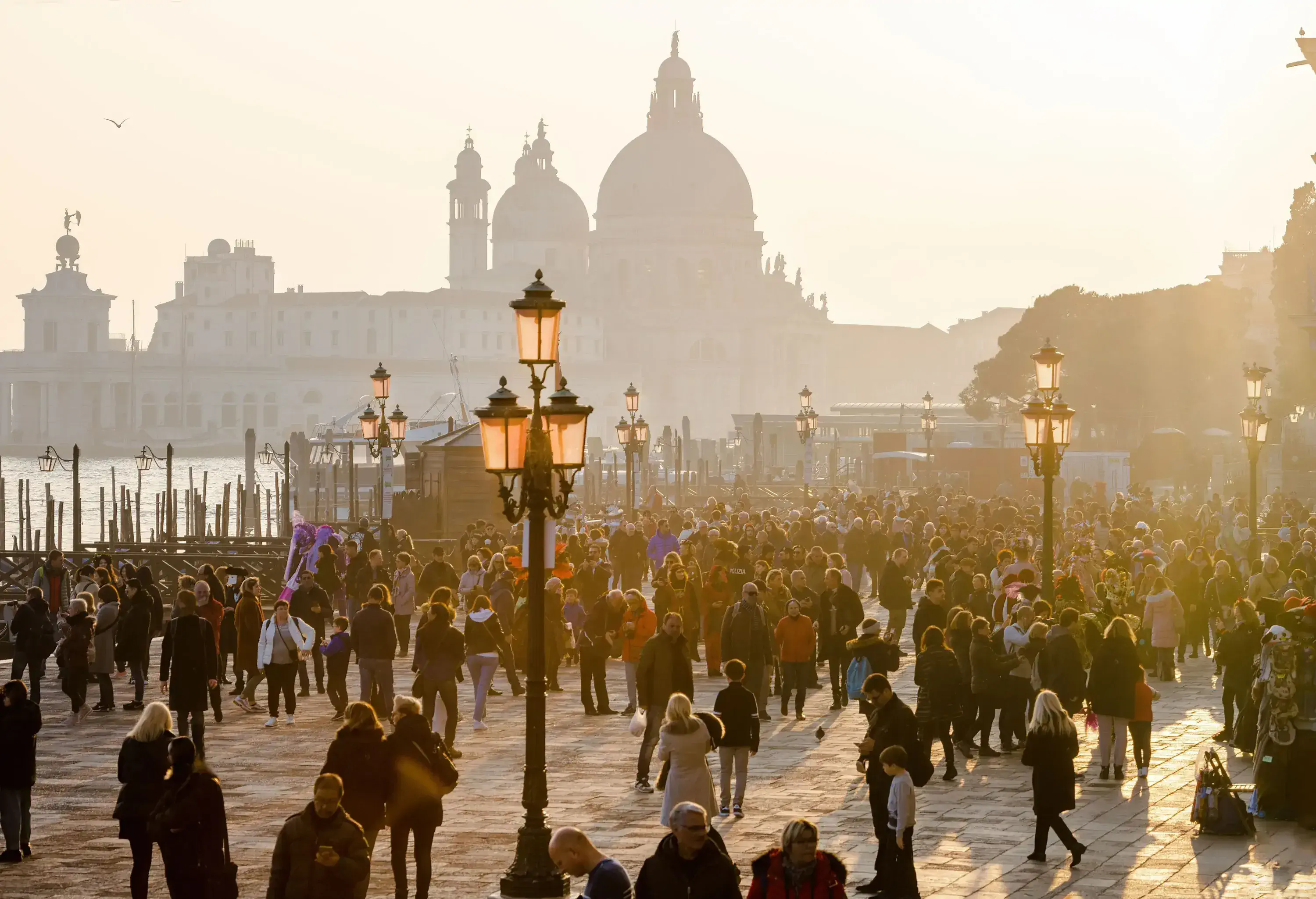 People fill a city square with ornate lampposts, against a backdrop of distant domed buildings under a hazy bright sky.