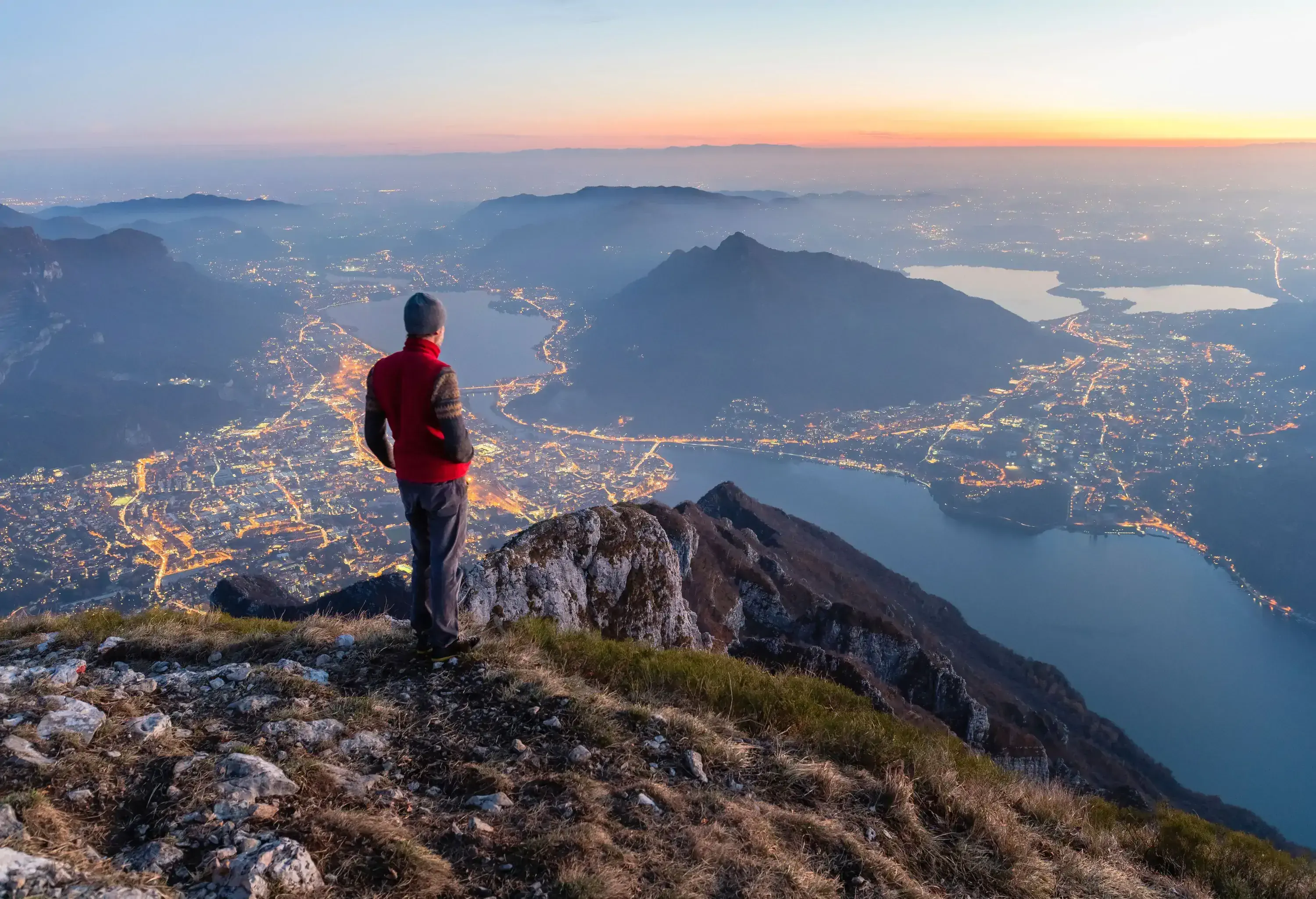 A man in a red jacket standing on a mountain top looking over a lake and illuminated cityscapes.