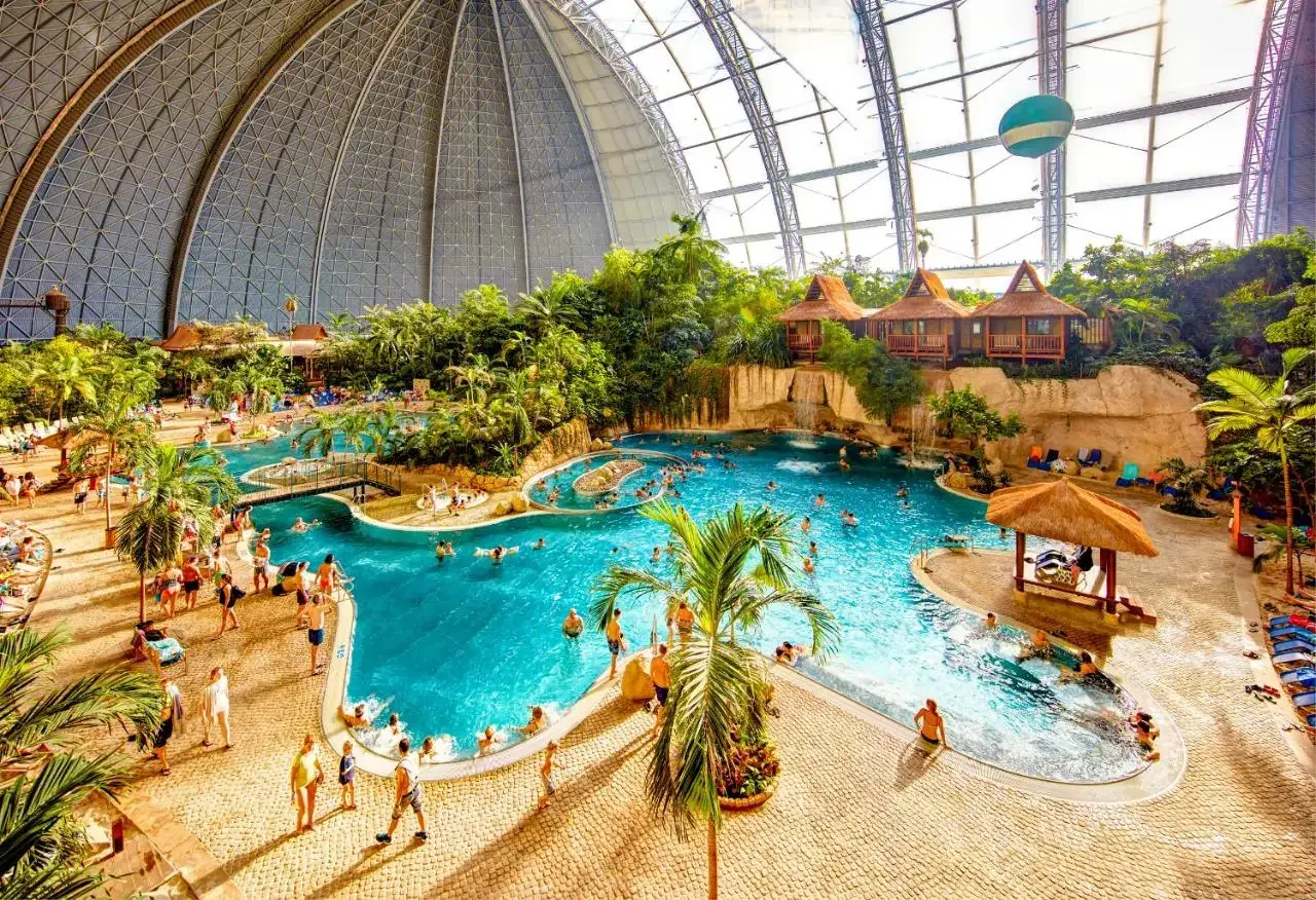 High angle view of a large indoor swimming pool with greenery and people built in a former Zeppelin hangar