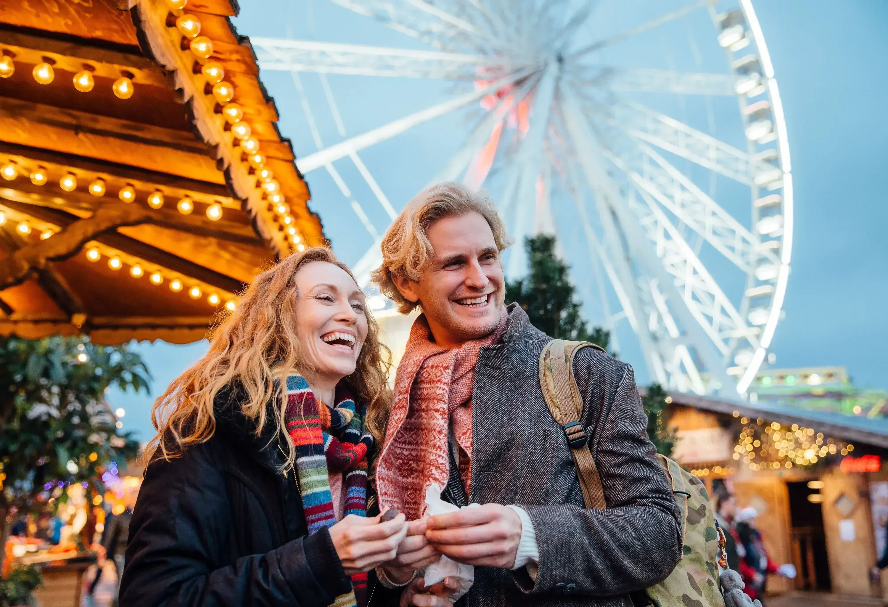 A smiling couple stands at an outdoor Christmas market, with a brightly lit Ferris wheel behind them.