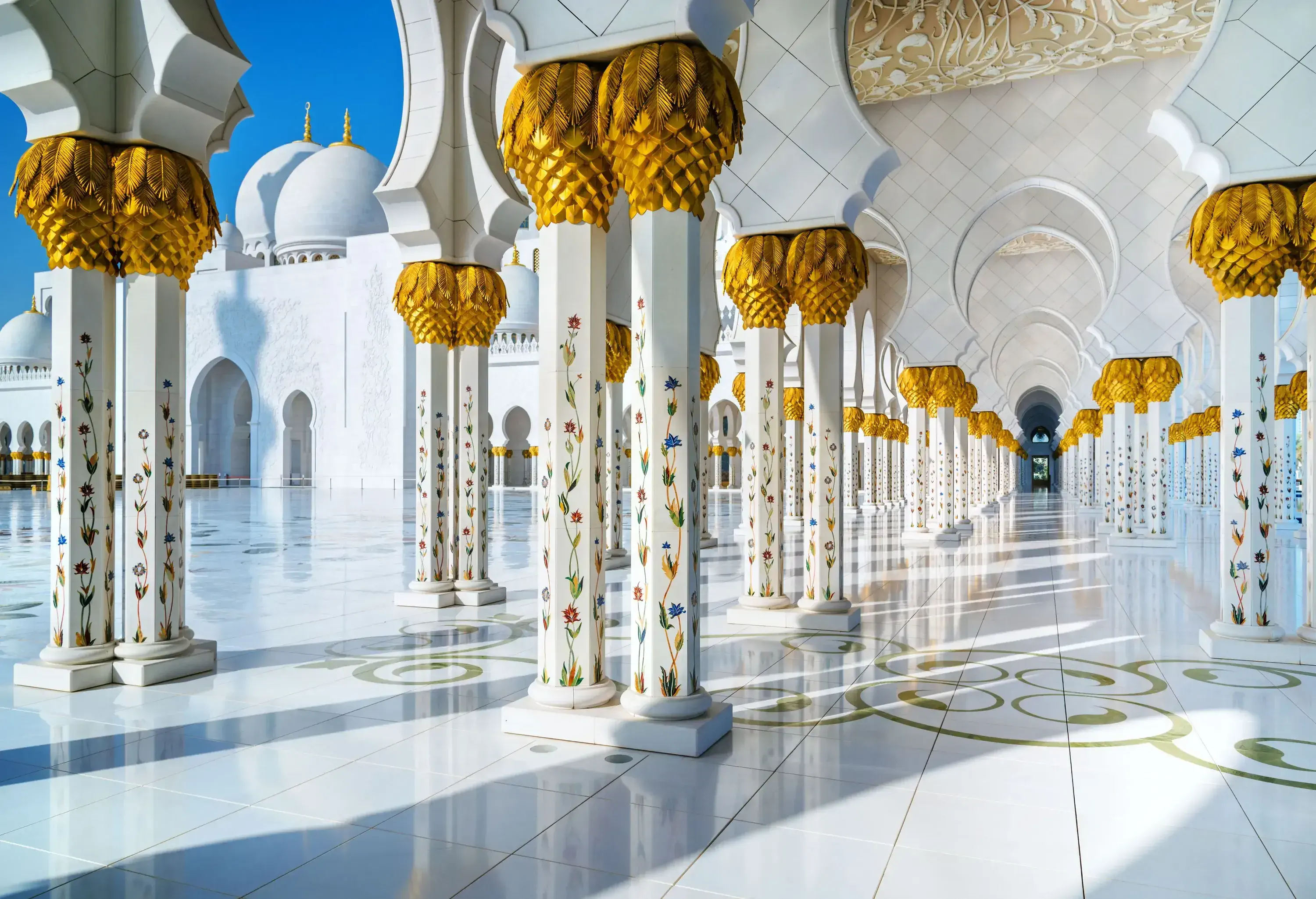 A grand white Mosque interior features intricate marble flooring adorned with floral designs and intricately designed pillars.