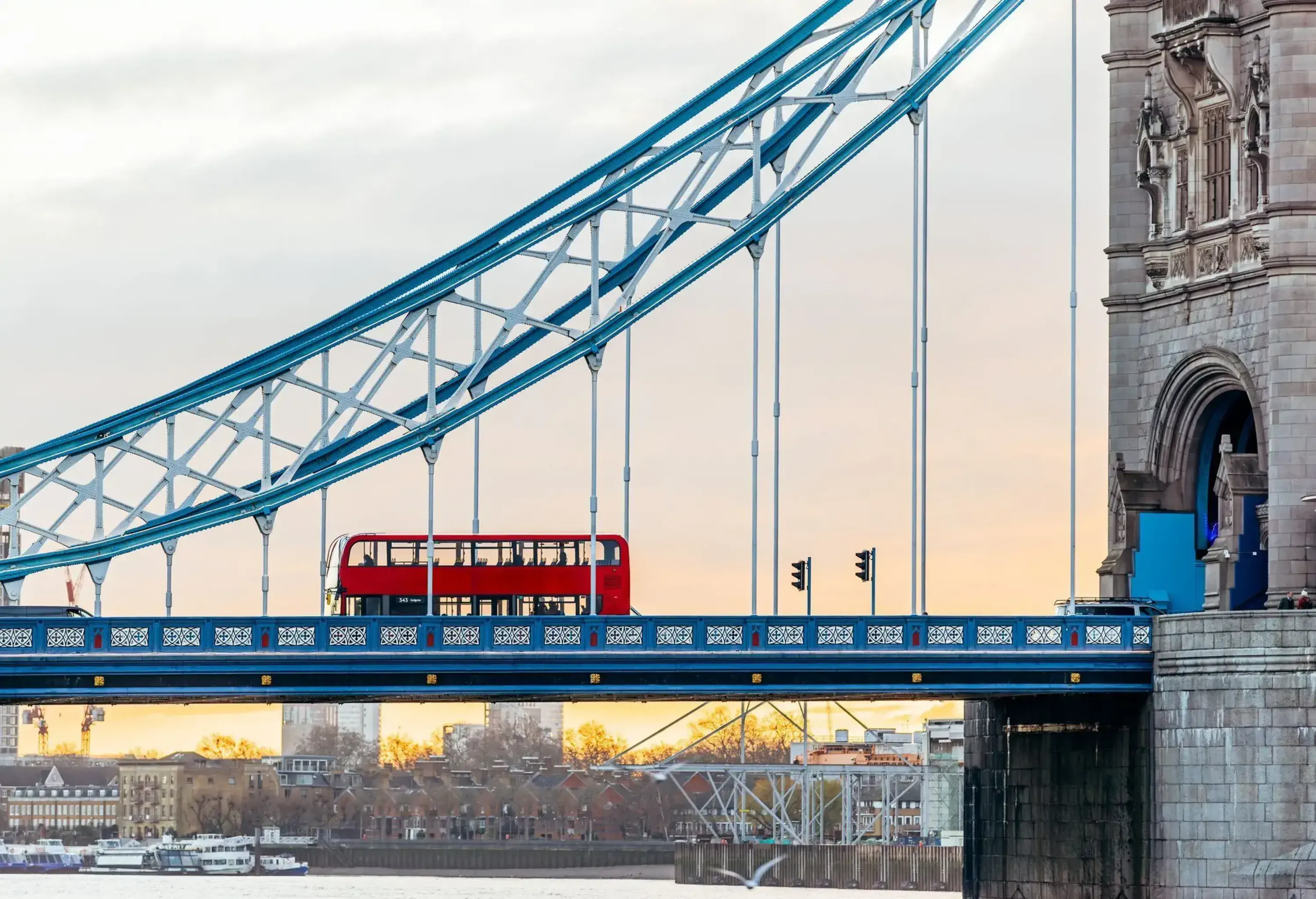 A red double-decker bus crosses Tower Bridge in London at sunset, with part of the bridge's structure and a stone tower visible.