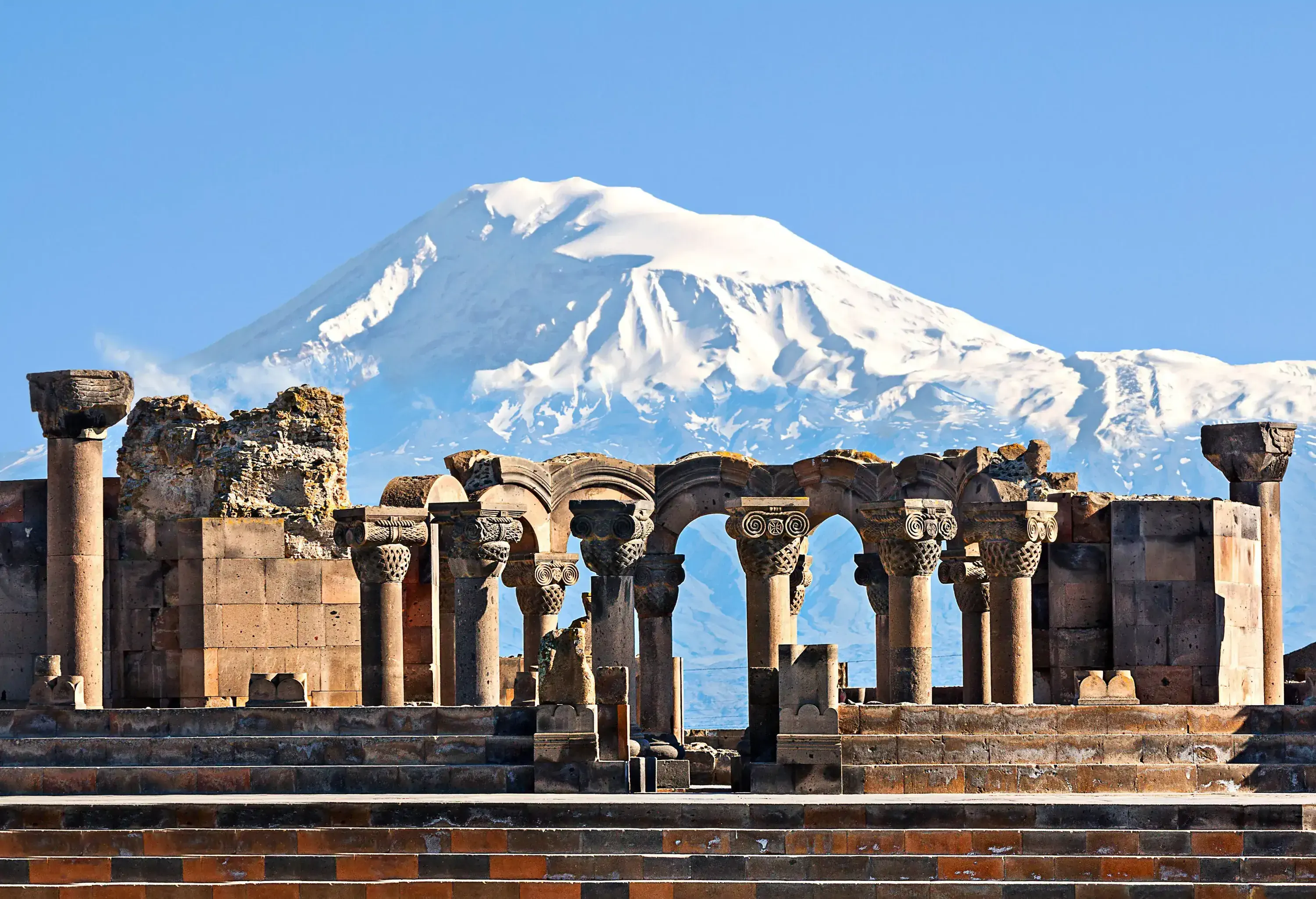 Ruins of a cathedral with the remains of ornate columns with a snow-covered mountain against the cloudless blue sky in the background. 
