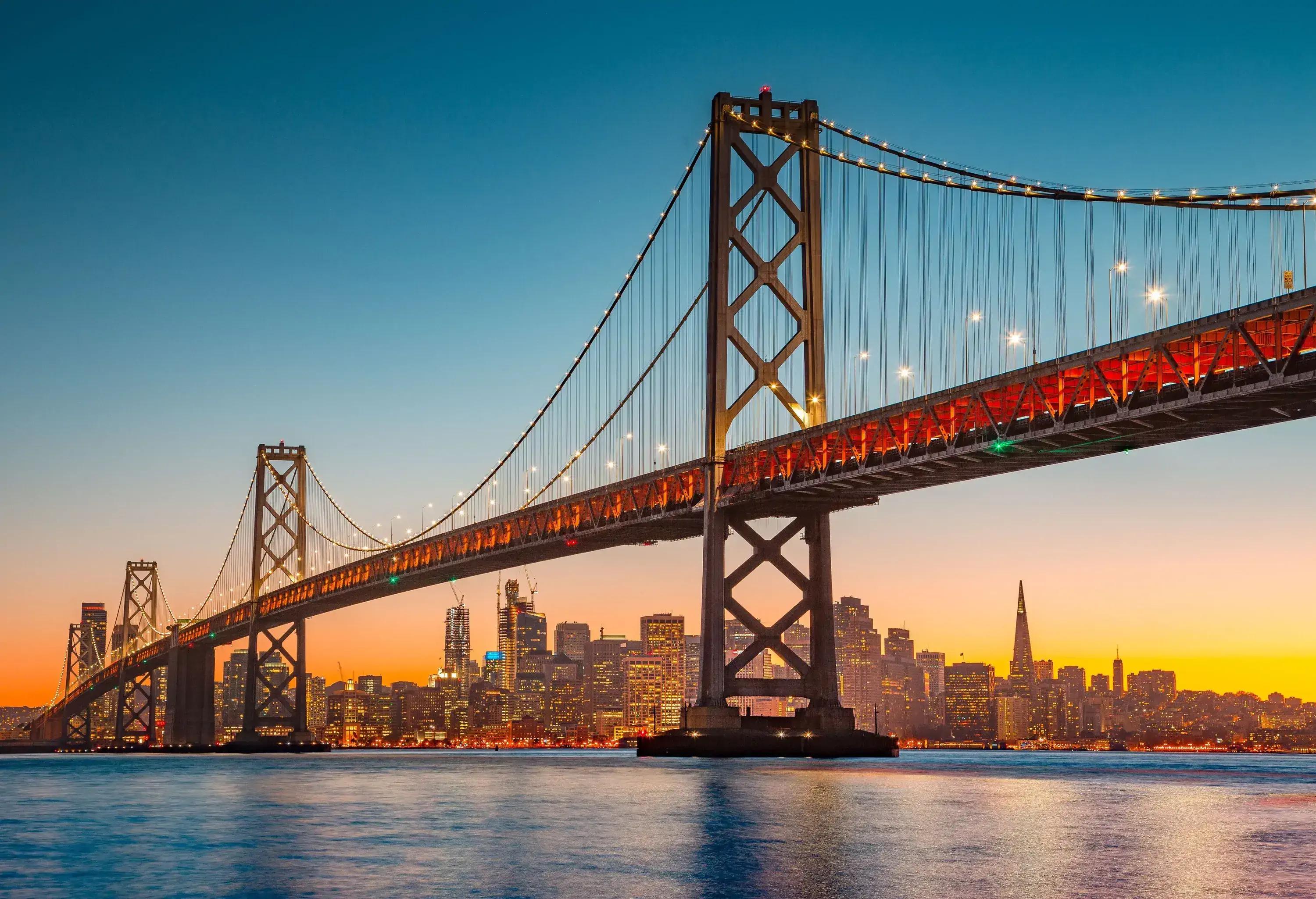 The Bay Bridge spanning the San Francisco Bay, with the lit city buildings in the background.