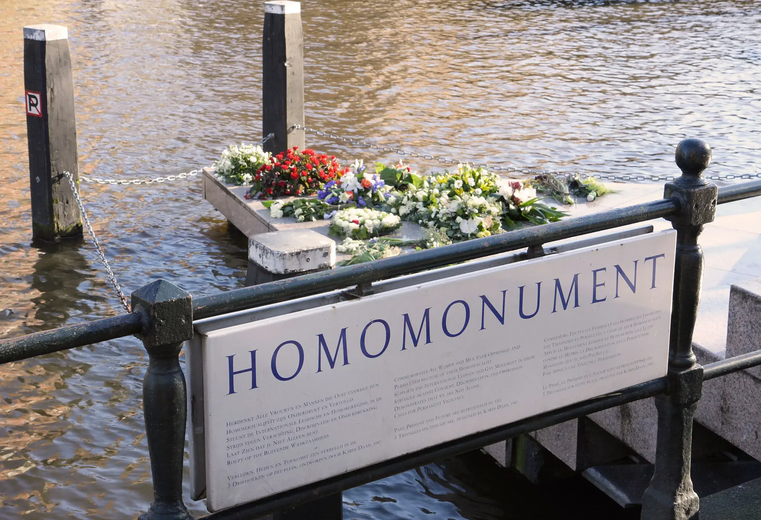 Colourful flowers laid in a triangular homomonument on the canal bank guarded by wooden posts and metal chains.