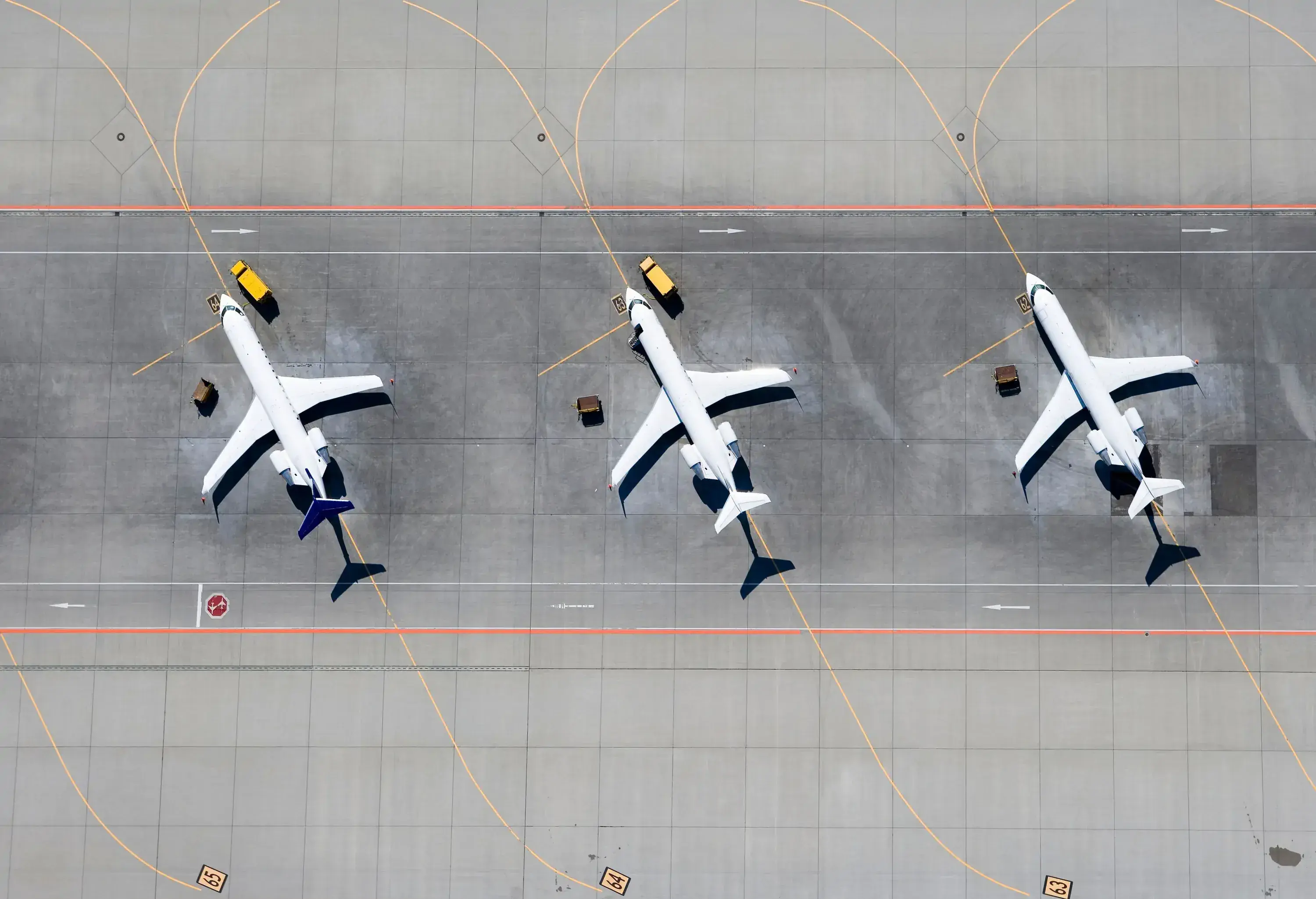 Aerial view of three parked airplanes on an airport tarmac, aligned in a row. Each plane is accompanied by small service vehicles.