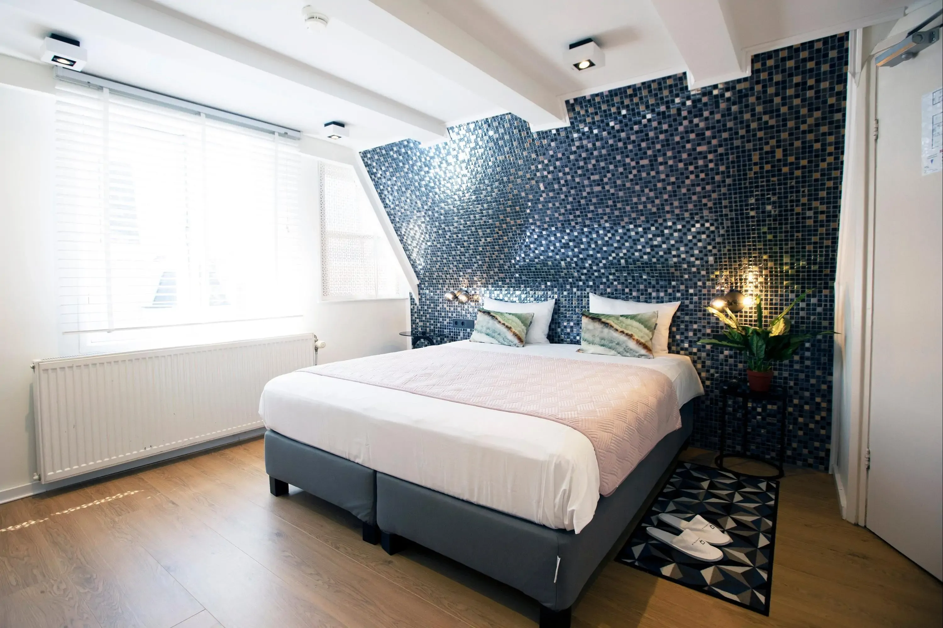 Hotel room with a modern, cozy design. Features a double bed with purple accents, a chaise lounge, desk, and a large window with purple curtains.