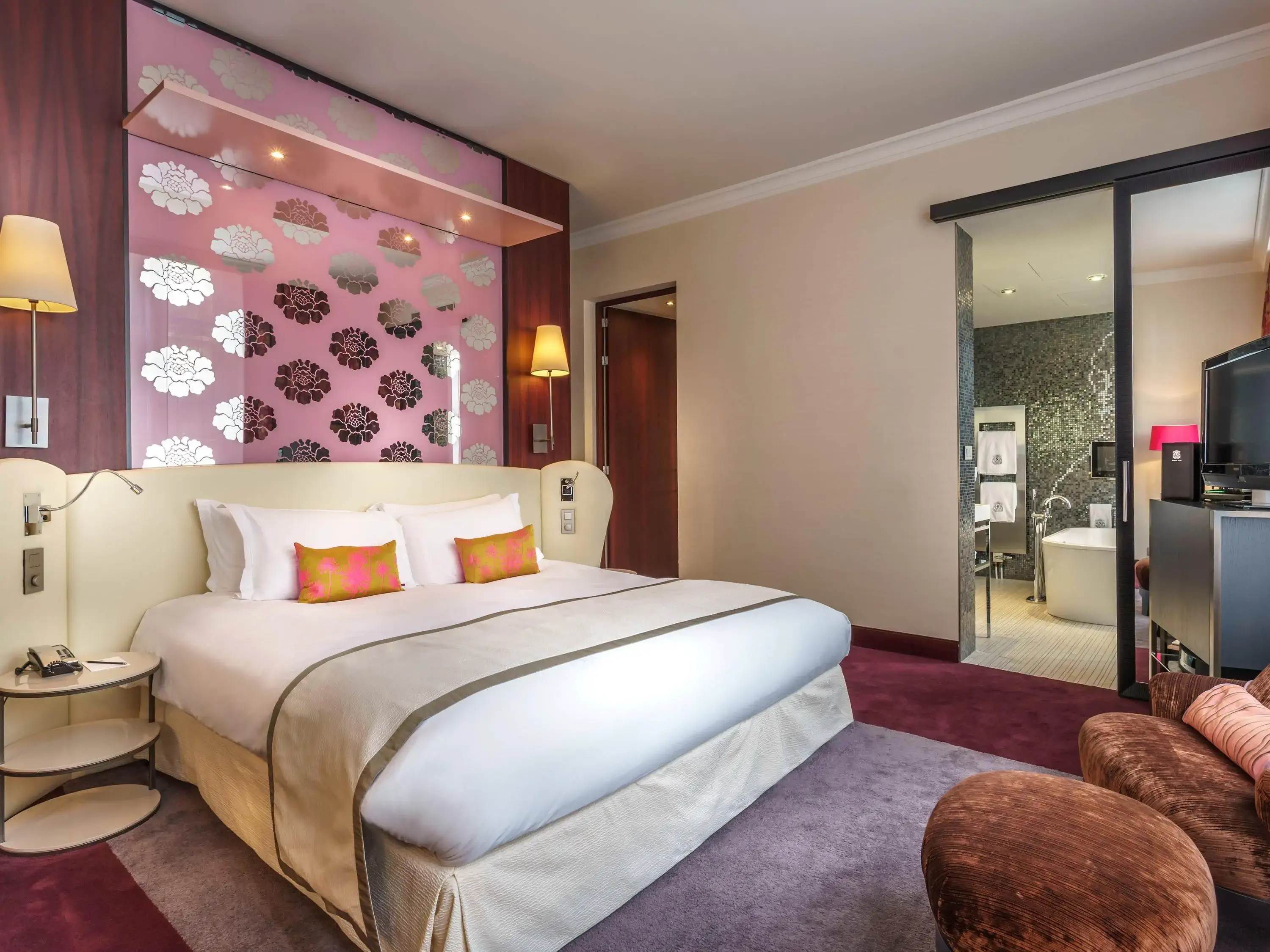 Spacious hotel room with a king-sized bed, floral-patterned wall, warm lighting, and modern decor. Open glass doors reveal a stylish bathroom.