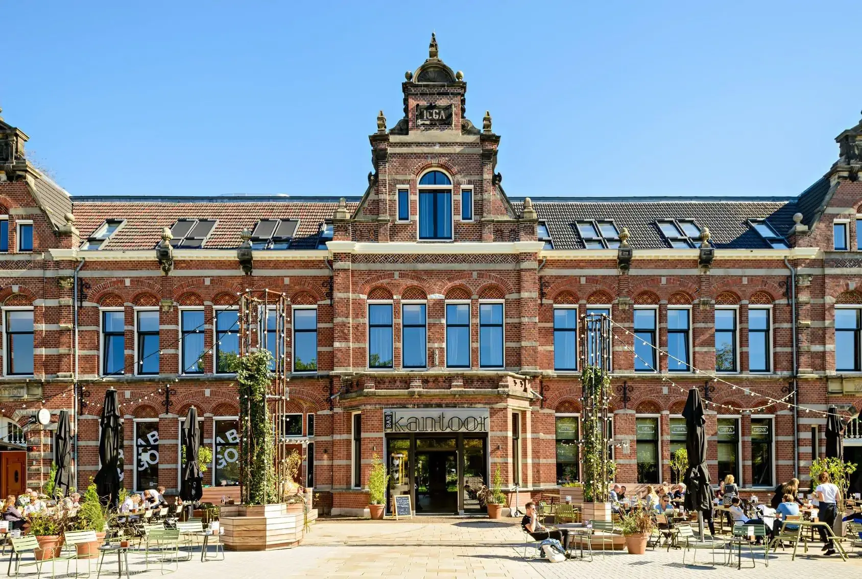 Historic brick building with large windows under a clear blue sky. Outdoor seating with people dining, creating a lively, inviting atmosphere.