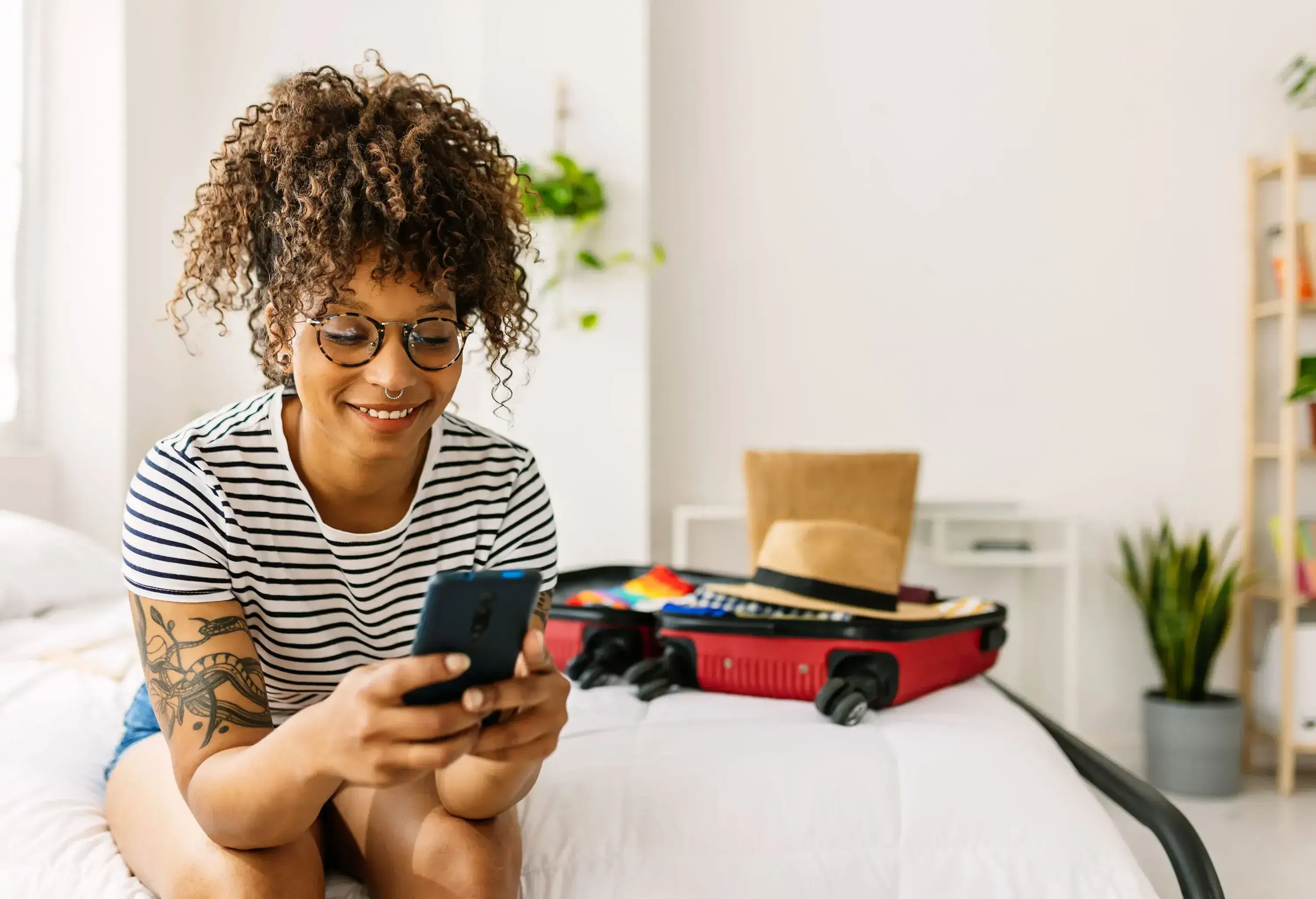 A young woman is sitting on a bed, smiling at her phone with an open suitcase next to her.