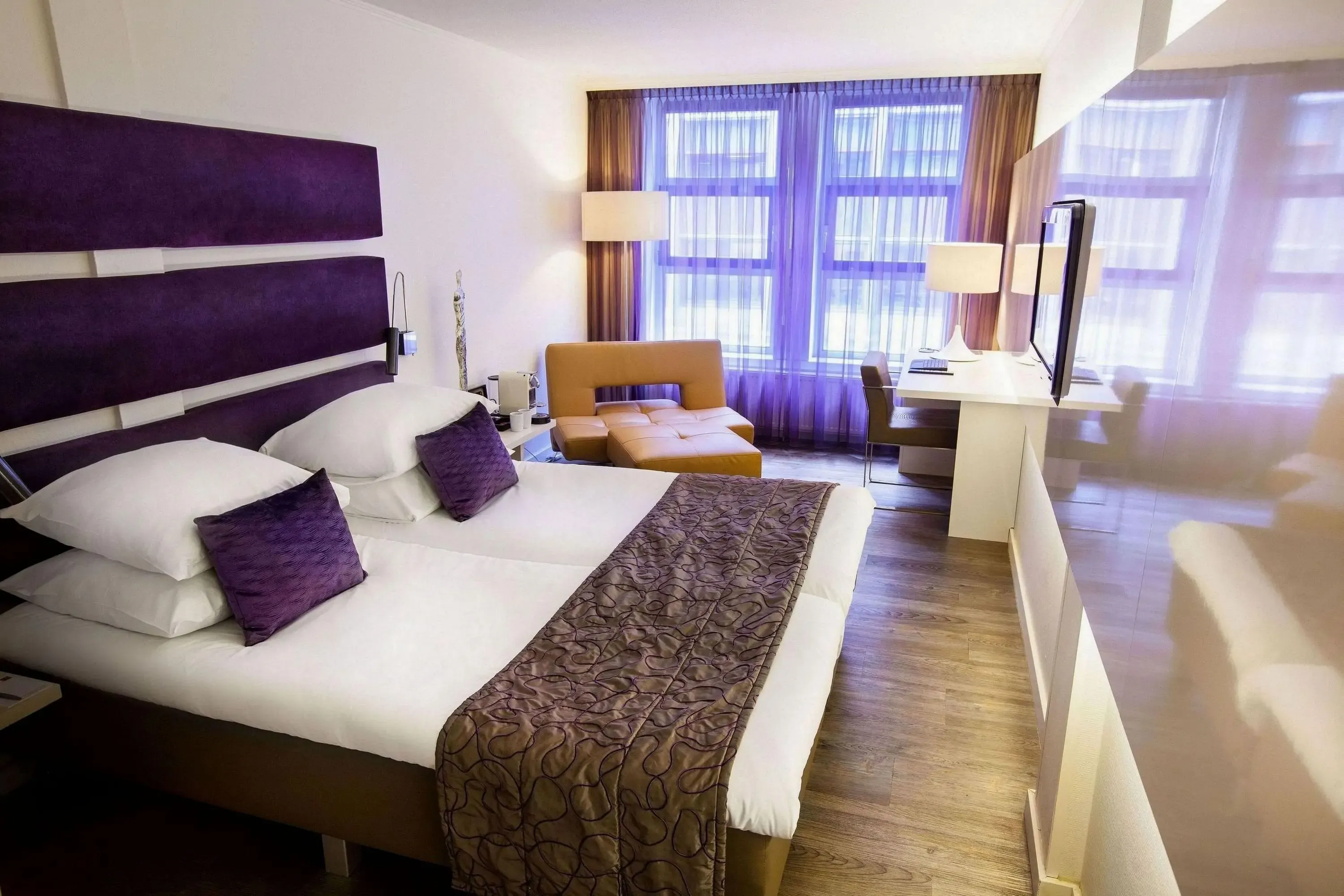 Hotel room with a modern, cozy design. Features a double bed with purple accents, a chaise lounge, desk, and a large window with purple curtains.