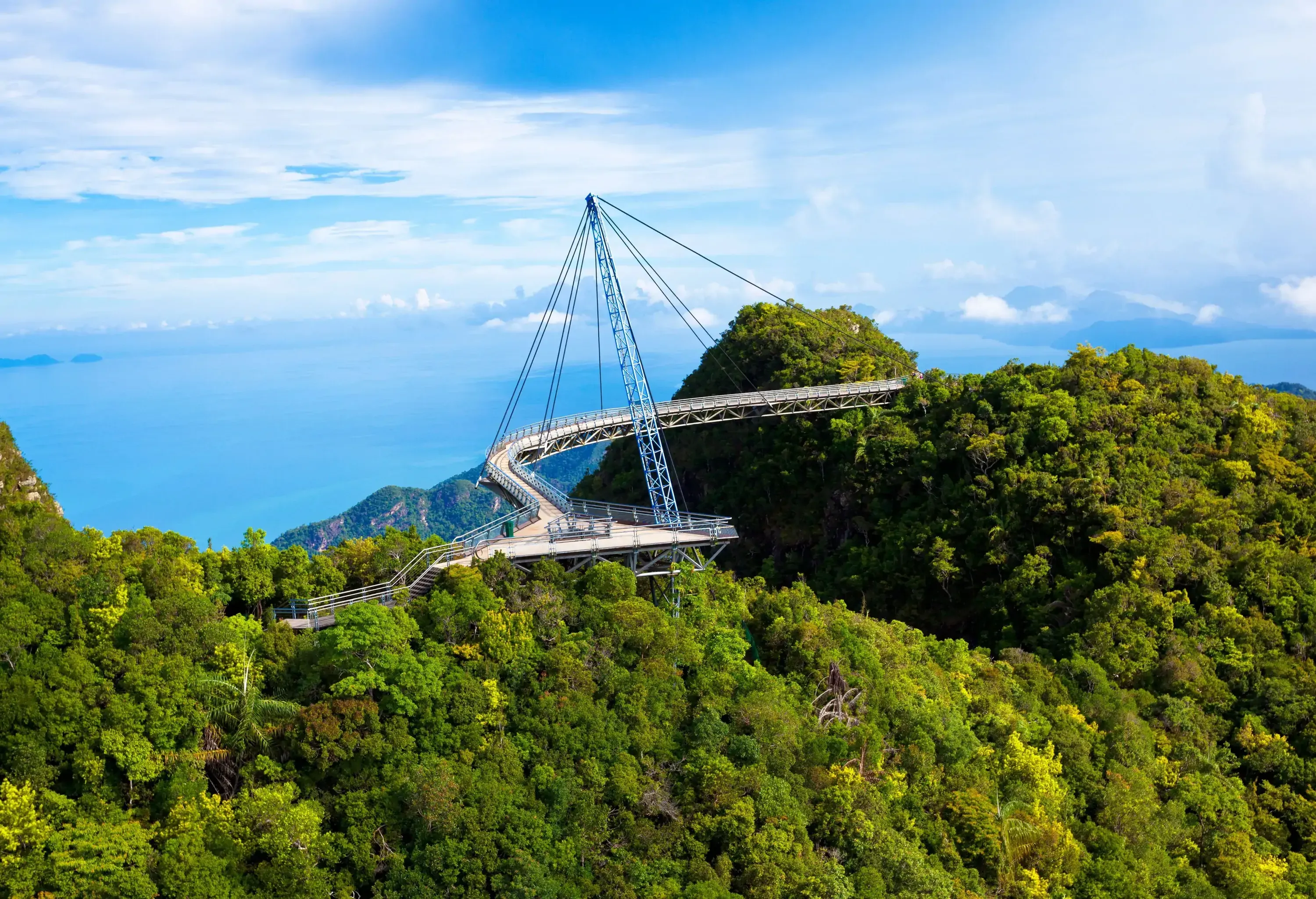 An impressive cable bridge over the tropical rainforest island surrounded by the sea against the cloudy blue sky.