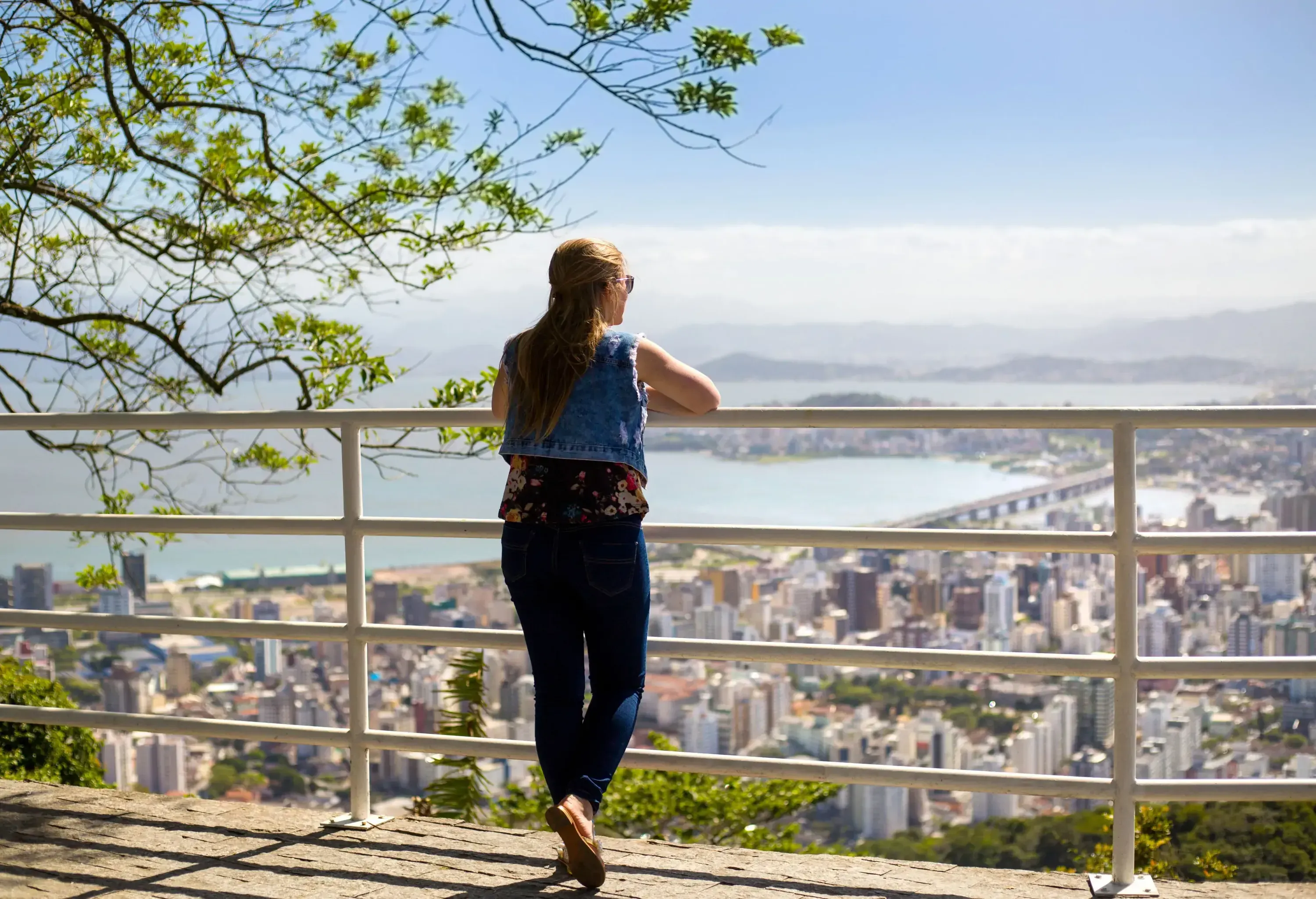 Back view of a blonde lady leaning on a white fence overlooking the compact buildings on the mainland sprawling to an island linked through a bridge. 