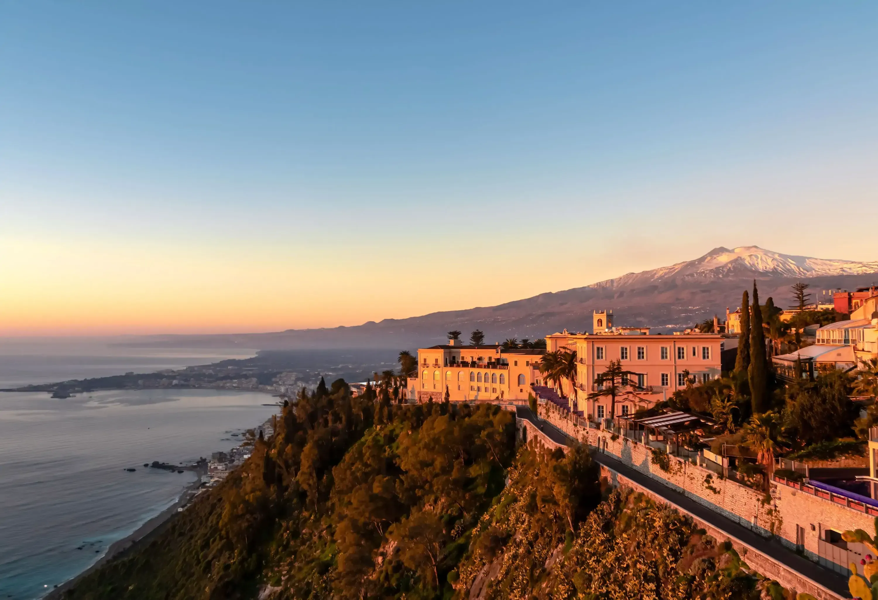 Sunset over a clifftop town, showcasing a historic palace-like building perched overlooking the sea, with a snow-capped mountain in the distance.