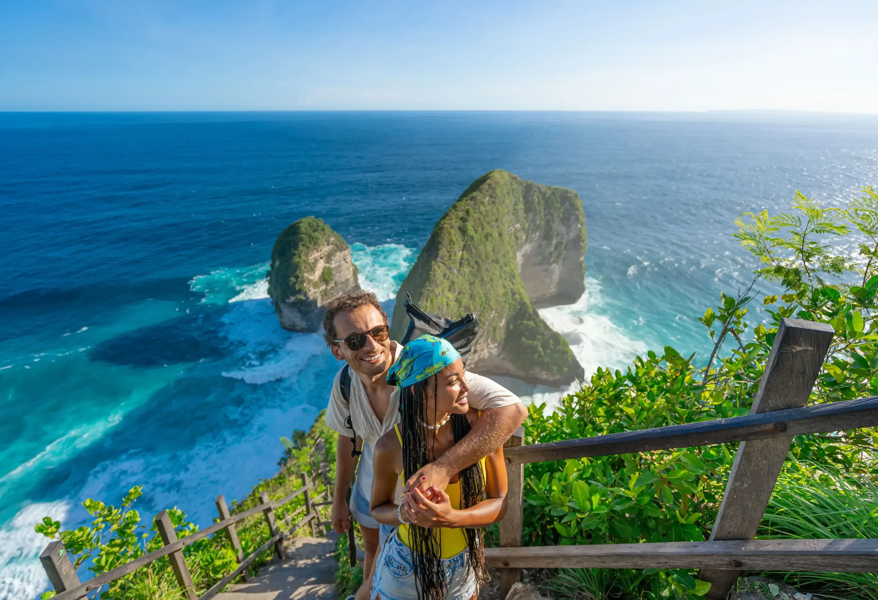 Family vacation lifestyle. Happy multiracial couple stand at viewpoint, looking at the beautiful beach under high cliff. Travel destination in Bali. Popular place to visit on Nusa Penida island.